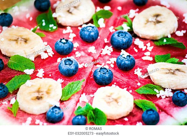 Sliced juicy watermelon pizza on wood, closeup view from above. Ingredients are watermelon, blueberries, banana, mint, and coconut shavings