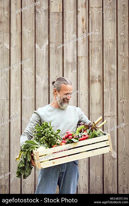 Bearded mature man carrying vegetable crate while standing against wall