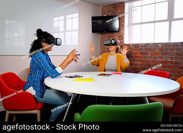 Diverse female colleagues sitting at work wearing virtual reality set and playing