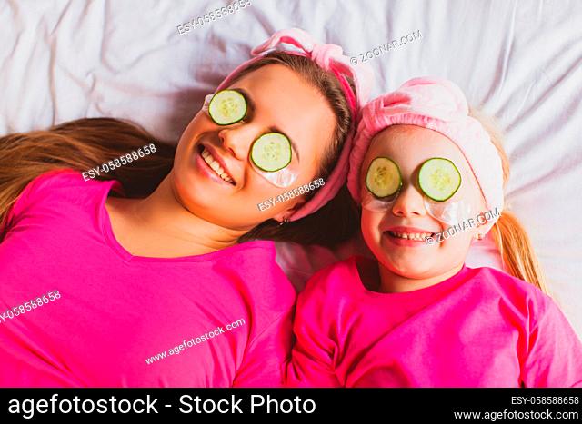 Close-up top view little girl and young woman faces with slices of cucumber on their eyes. Portrait of mother and daughter doing spa procedures at home during...