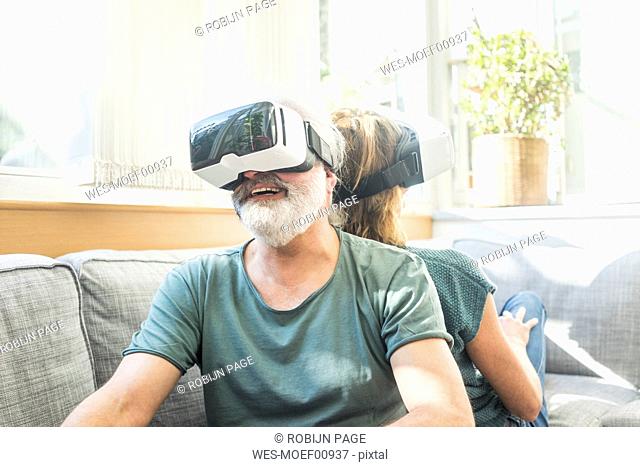 Happy mature couple sitting on couch at home wearing VR glasses