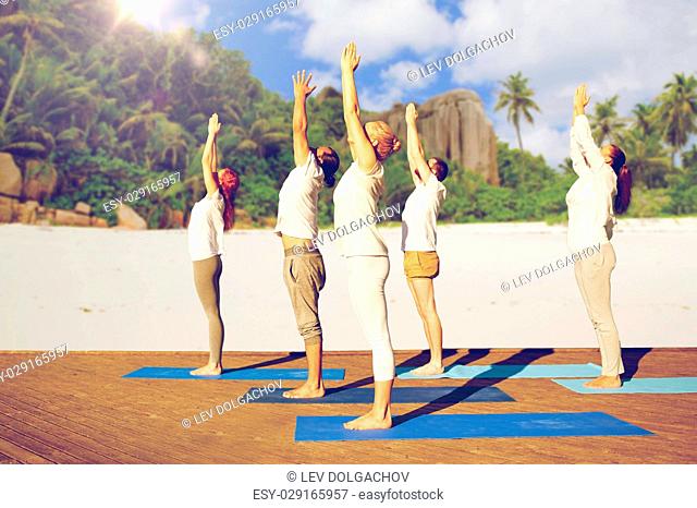 fitness, sport, yoga and healthy lifestyle concept - group of people making upward salute pose over tropical beach background