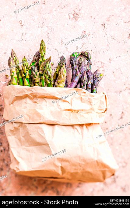Top view of bunch of fresh purple and green asparagus spears on the pink marble table in modern kitchen. Copy space on the paper bag