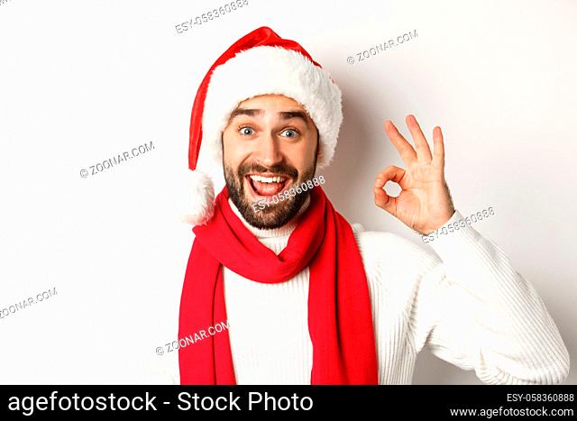 New Year party and winter holidays concept. Close-up of happy attractive man in santa hat showing ok sign, celebrating christmas, white background