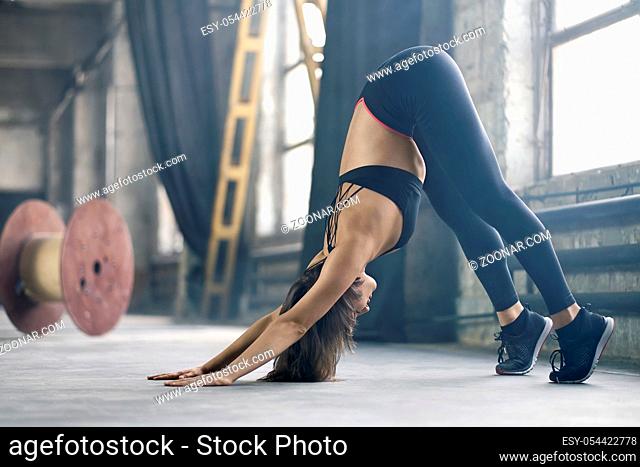 Sportive girl with closed eyes in a black sportswear engaged in yoga on the windows background in the loft style hall. She does a downward facing dog asana on...