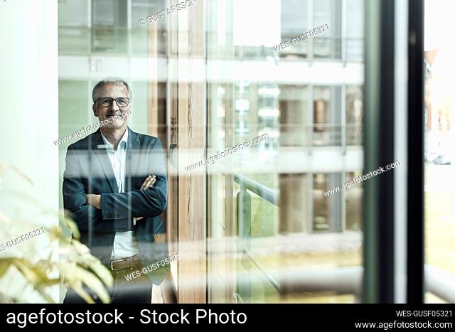 Smiling businessman with arms crossed seen through glass in office corridor