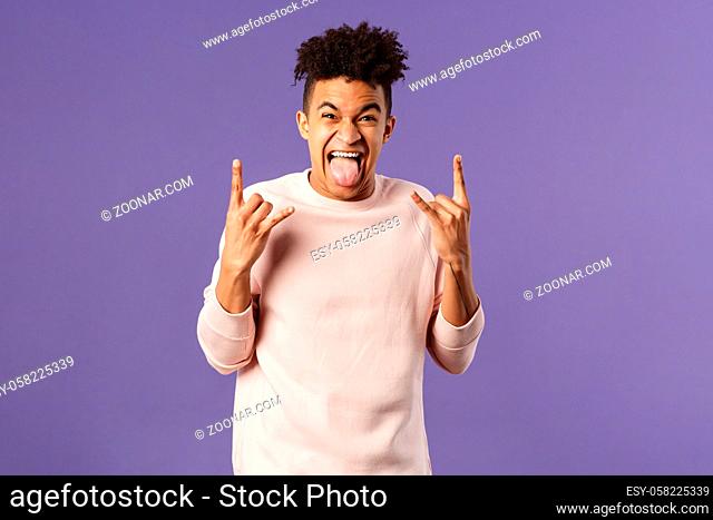 Portrait of enthusiastic, excited funny young man having fun, enjoying awesome rock concert, show heavy metal sign, stick tongue and rejoicing hear favorite...