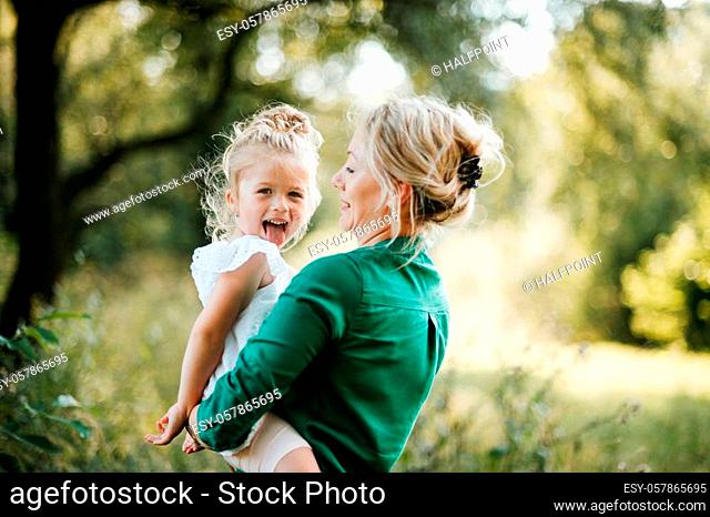 Beautiful young mother in green sunny summer nature holding her cute small daughter in the arms, having fun. Copy space