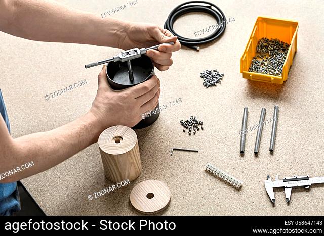 Man with tattoo is using a t-wrench on a black metal cylinder on the light table. On the table there are wooden billets, cable, plastic box with screws, tools