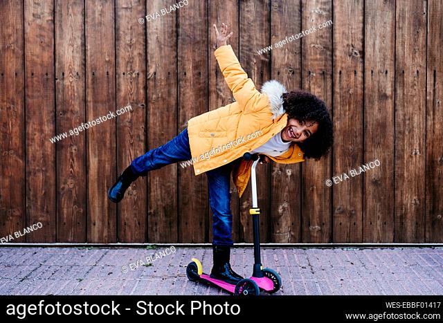 Playful girl riding push scooter against wooden wall