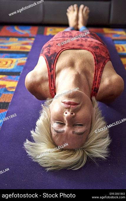 A woman, 60+ years old, lies on a mat on the floor with her arms under her arched back while she practices yoga in her apartment home, Windsor, Ontario, Canada