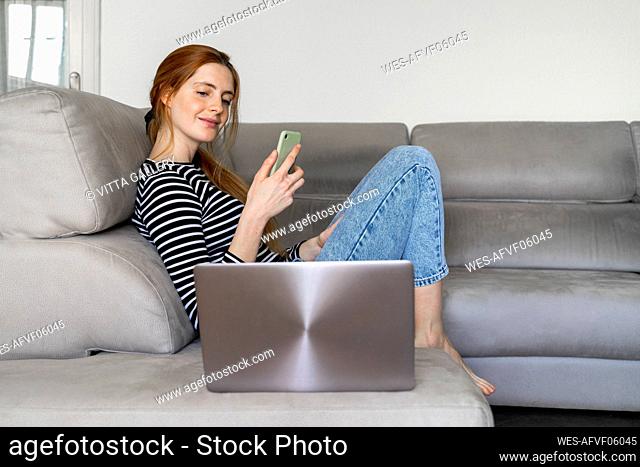 Young woman sitting on couch, using smartphone and laptop