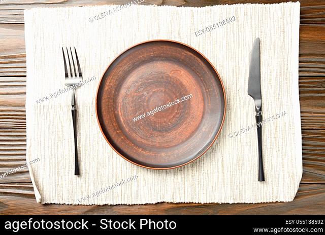 Top view at empty clay plate knife and fork aside on grey napkin on brown wooden kitchen table