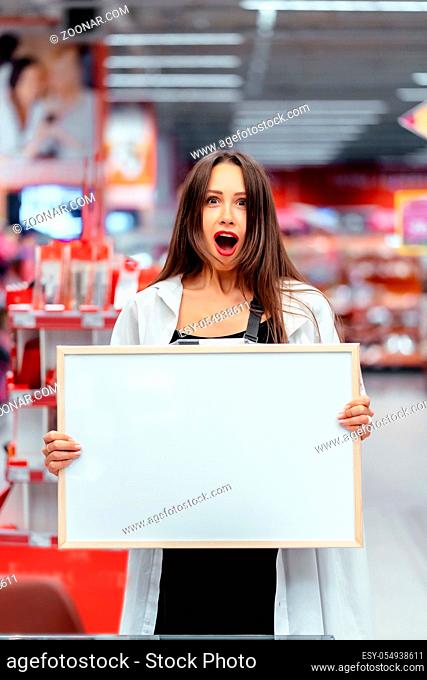 Smiling brunette woman holding white blank board in supermarket blur background
