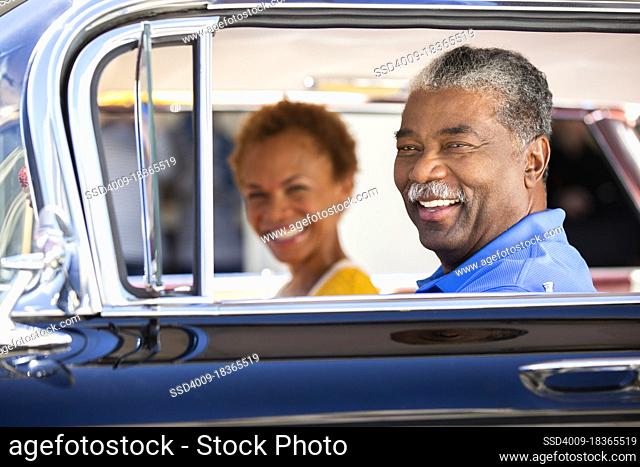 Older couple sitting in a classic car looking to camera smiling