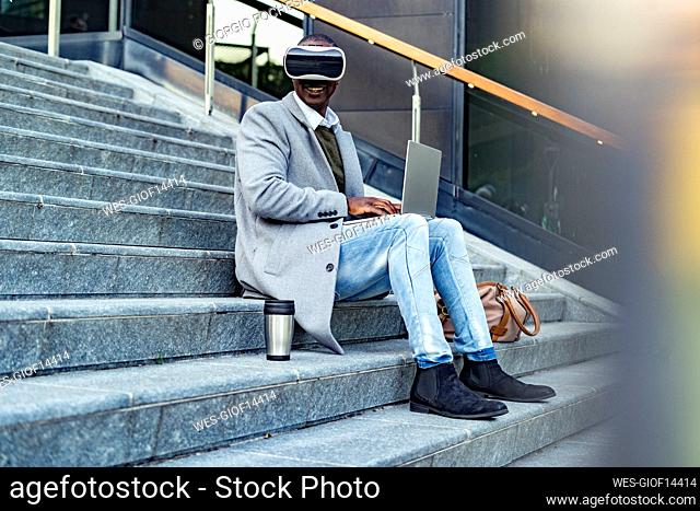 Businessman with virtual reality headset using laptop sitting on steps