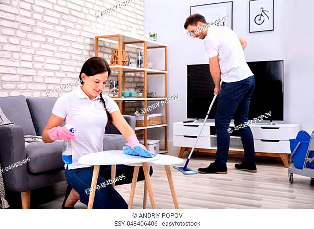 Two Smiling Young Workers Cleaning The Table And Mopping Floor In The Living Room