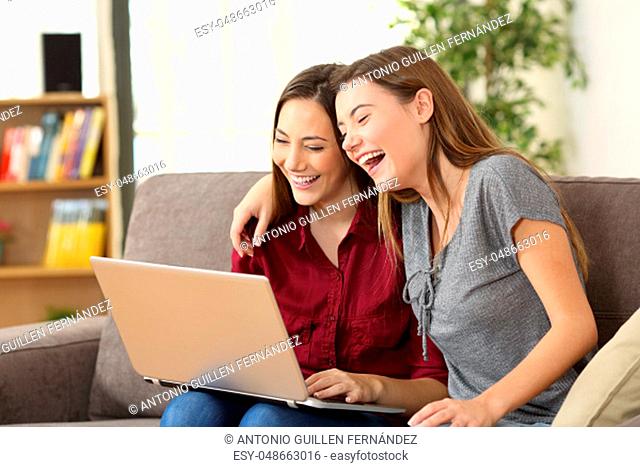 Two playful roommates watching videos on line together in a laptop sitting on a couch in the living room at home