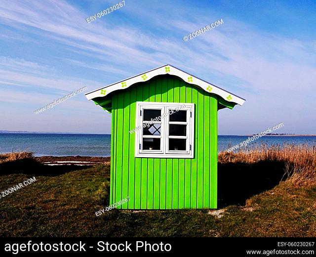 A sweet little wooden beach hut, painted bright green with white roof and window frame against background of sea and a windswept blue sky