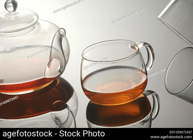 Transparent glass teapot and cup of tea on the reflective surface on a light gray background