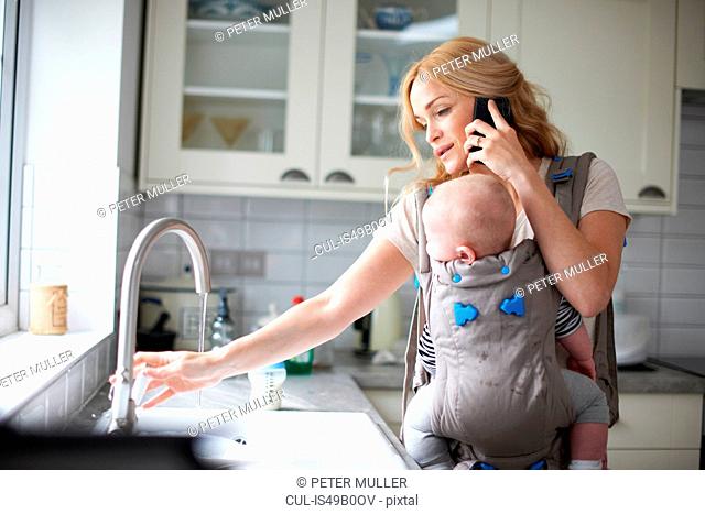 Woman holding baby boy in sling, using smartphone, turning on tap to do washing up