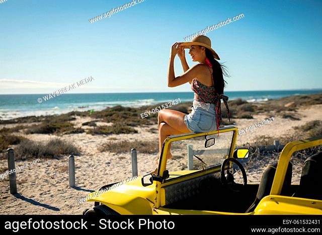 Happy caucasian woman sitting on beach buggy by the sea wearing straw hat looking toward sea