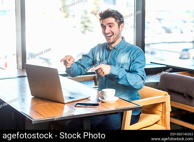 Yes, it`s you! Side view portrait of happy handsome bearded young freelancer in blue jeans shirt are sitting in cafe and making video call on laptop