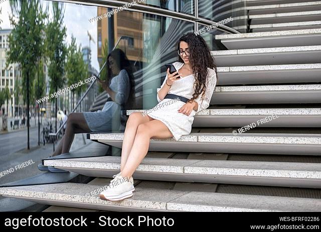 Young woman with long hair using mobile phone while sitting on steps in city