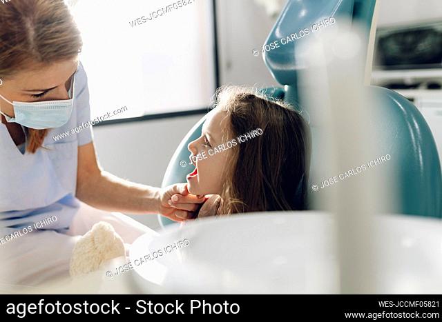 Dentist wearing protective face mask examining teeth of little girl at clinic