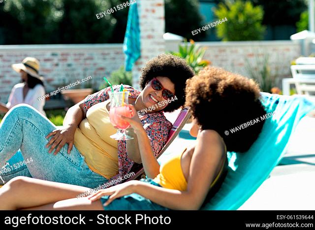 Biracial female friends with afro hair toasting cocktails while lying on deckchairs at pool party