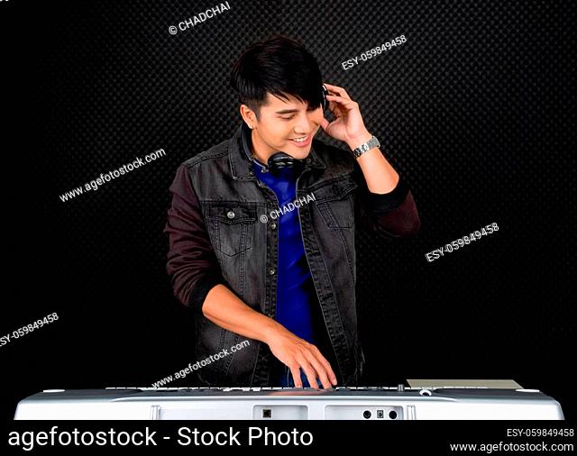 Young asian man with headphones playing an electric keyboard in front of black soundproofing walls. Musicians producing music in professional recording studio