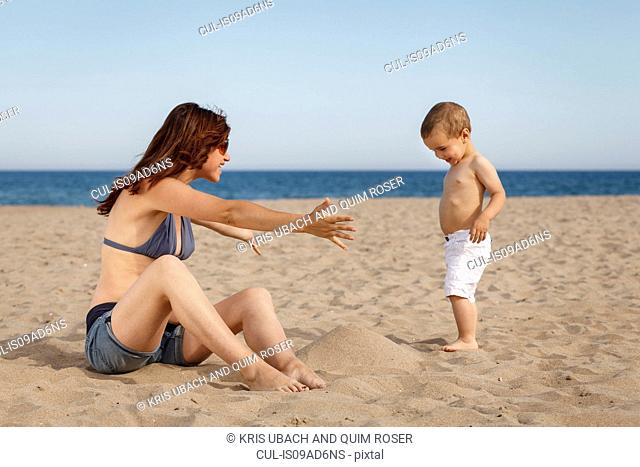 Pregnant woman sitting on beach with arms open to toddler