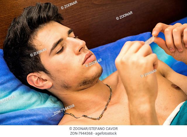 Young handsome man in bed with a flu or measuring fever with thermometer in his hand, checking the temperature