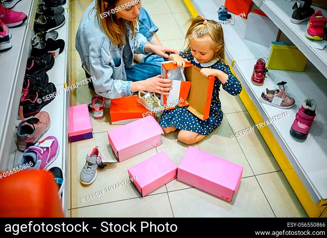 Mother and little baby opens box with shoes in kid's store. Mom and adorable girl near the showcase in children's shop, happy childhood