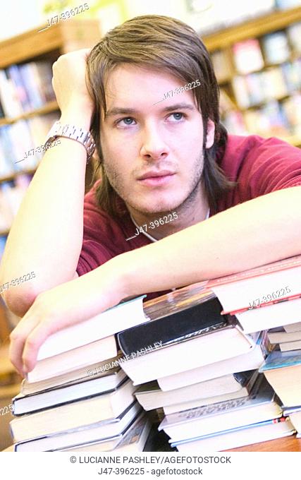 24 year old man leaning over a pile of books, head in hand, in the library