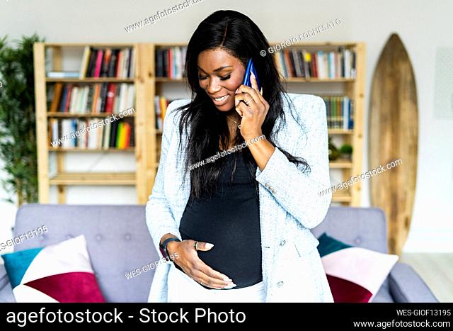 Smiling pregnant female freelancer touching abdomen while talking on mobile phone at home