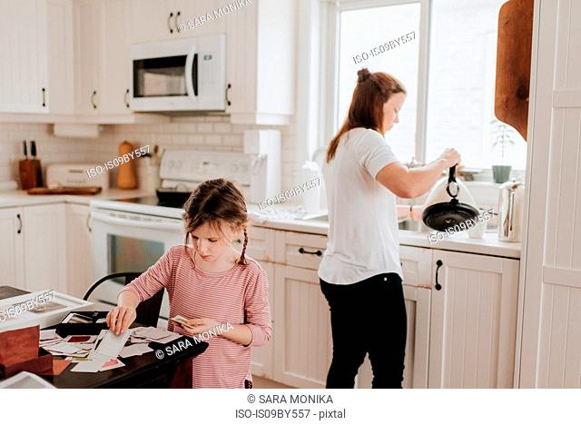 Girl arranging cards in kitchen, mother pouring hot water in background