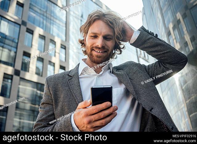 Smiling businessman with hand in hair using mobile phone against office building at downtown