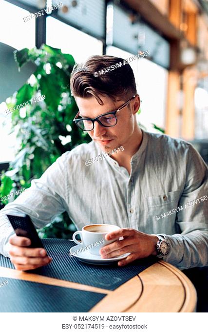 Successful thoughtful young businessman in eyeglasses, using mobile phone, drinking coffee at cafe. Dressed in shirt