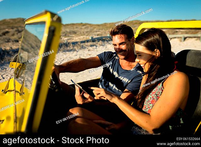 Happy caucasian couple sitting in beach buggy by the sea using smartphone