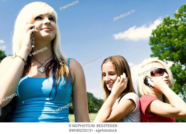 three young girls standing in a meadow phoning with a mobile with a smile