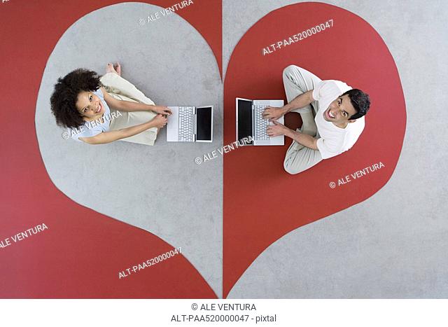 Man and woman sitting on large heart, using laptop computers, smiling up at camera