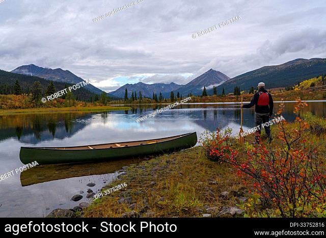 Man standing with his canoe on the shores of Rose Lake, Yukon in autumn.; Yukon, Canada