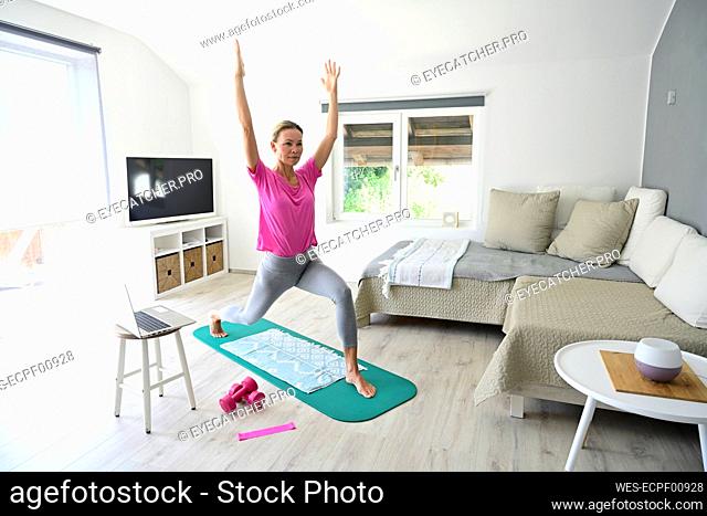 Mature woman with laptop practicing yoga on gym mat in living room