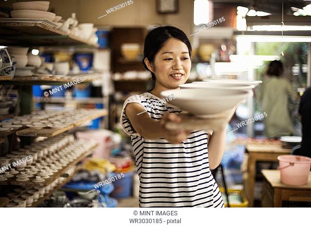 Woman standing in a workshop, carrying long wooden tray with porcelain bowls