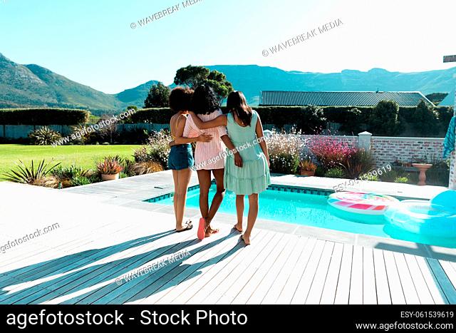 Biracial young female friends with arms around hanging out at poolside during summer, copy space