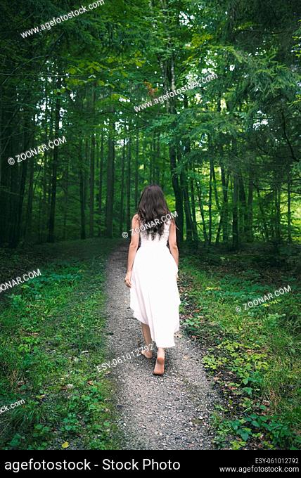 Back view of a young brunette woman, dressed in an elegant white dress, walking on a pathway through spring forest, with bare feet