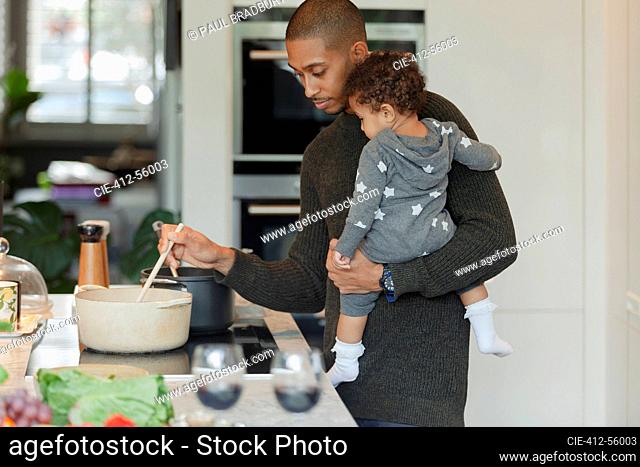 Father holding baby daughter and cooking dinner at kitchen stove