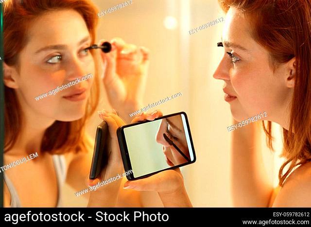 Attentive woman putting on make up watching video tutorial looking at mirror in the bathroom at home