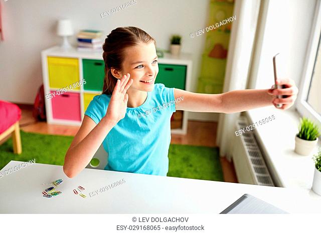people, children and technology concept - girl with laptop computer and smartphone taking selfie at home and waving hand